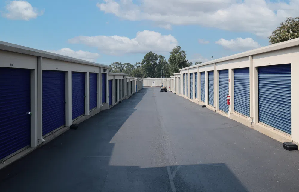 Outdoor view of a storage facility with rows of beige units and blue roll-up doors under a partly cloudy sky.