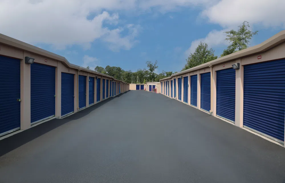 Wide view of a storage facility with rows of beige units featuring blue roll-up doors under a partly cloudy sky.