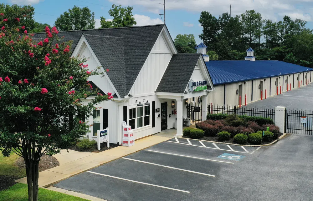 Midgard self storage facility with white office building, blue roof units, gated entrance, and parking lot under blue sky.