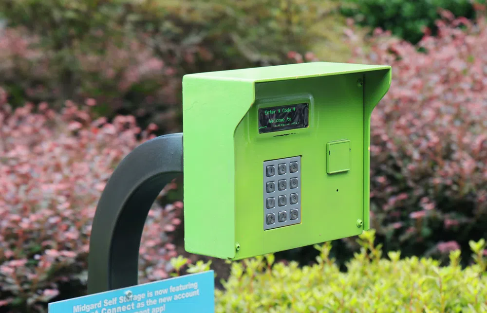 Green keypad security access system mounted on black armature with blurred shrubs background.