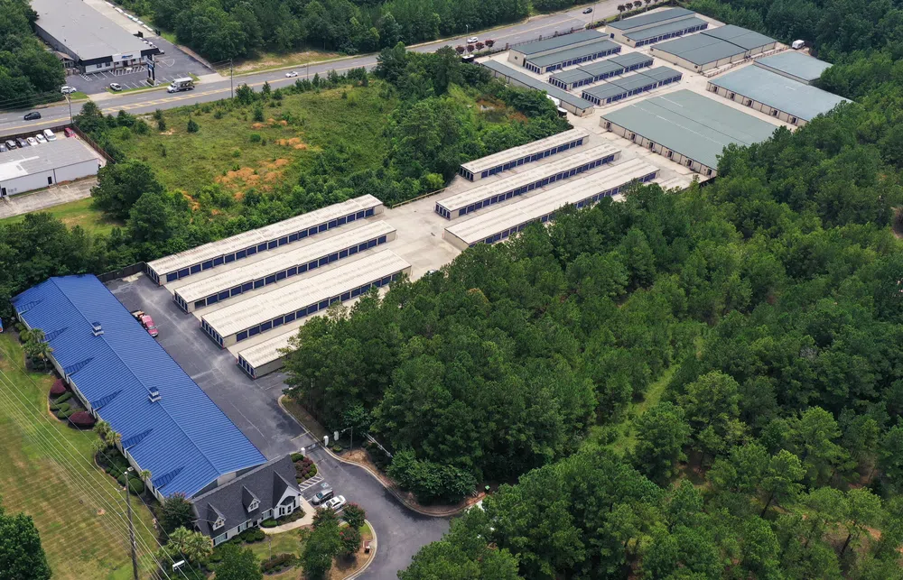 Aerial view of a self storage facility with rows of storage units surrounded by trees and nearby roads.