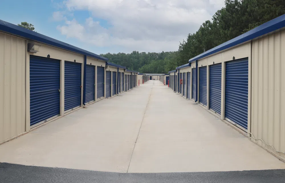 Outdoor storage facility with rows of beige units featuring blue roll-up doors under a partly cloudy sky.
