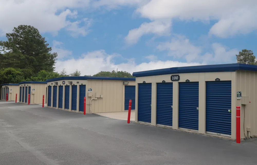 Row of beige storage units with blue roll-up doors and red bollards under a partly cloudy sky.