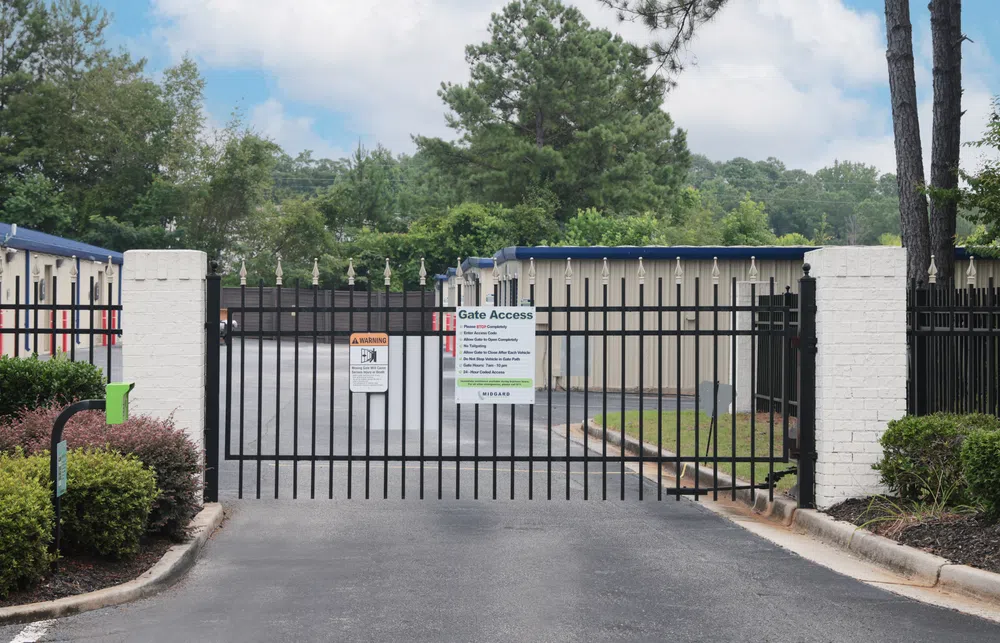 Black metal security gate with access instructions at entrance to storage facility with beige units and green trees.