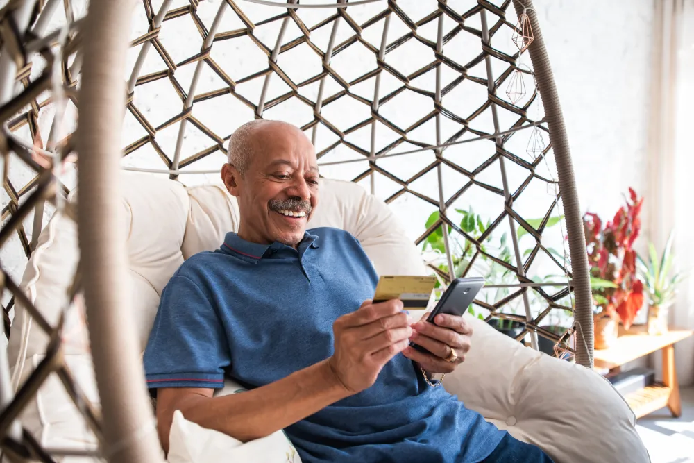 Smiling man sitting in a hanging chair holding a credit card and smartphone for online shopping at home.