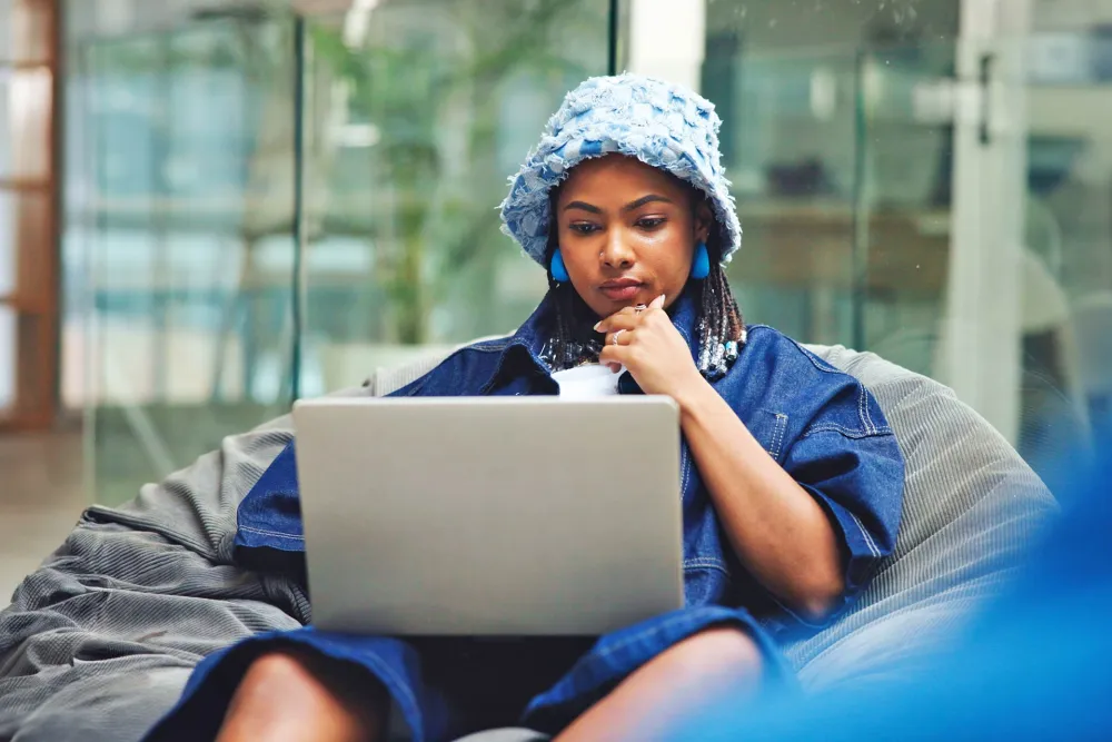 Young woman wearing a blue outfit and hat working thoughtfully on a laptop while sitting on a bean bag.