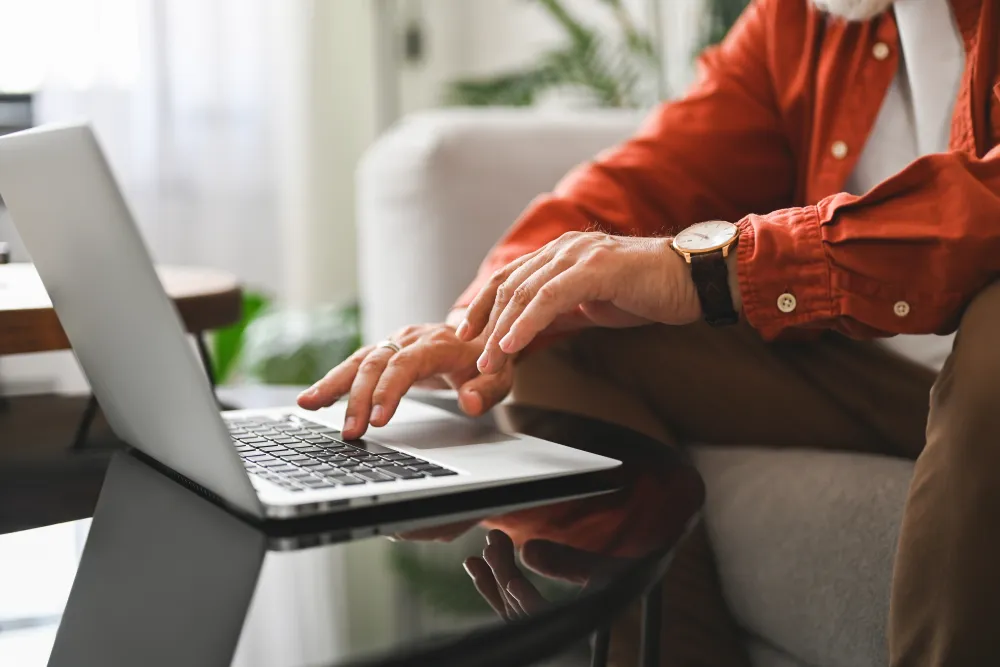 Close-up of a person wearing a watch typing on a laptop keyboard at a reflective glass table indoors.