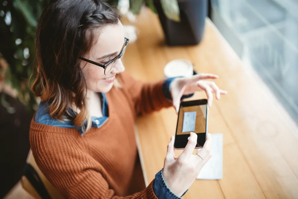 Woman in glasses taking a photo of a check using a smartphone at a wooden table by a window.