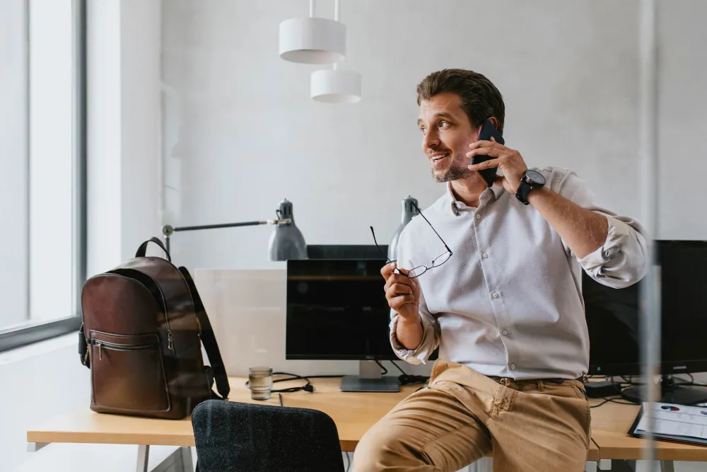 Man in white shirt sitting on desk holding glasses and talking on smartphone in modern office workspace