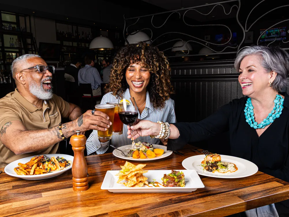 Three diverse adults toasting drinks at a restaurant table with gourmet food and smiles