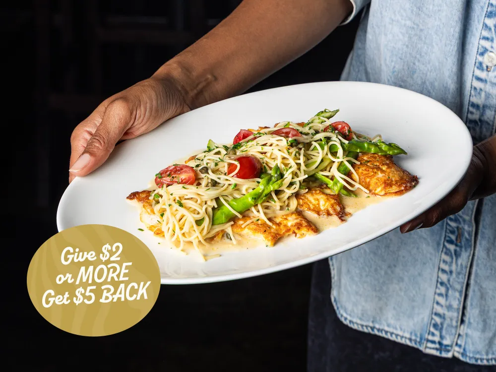 Hand holding a white plate with chicken breast topped with pasta, asparagus, cherry tomatoes, and herbs.