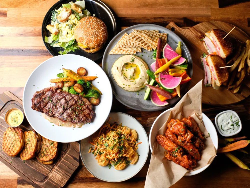 Assortment of grilled steak, chicken wings, sandwiches, hummus, salad, fries, and grilled bread on wooden table.