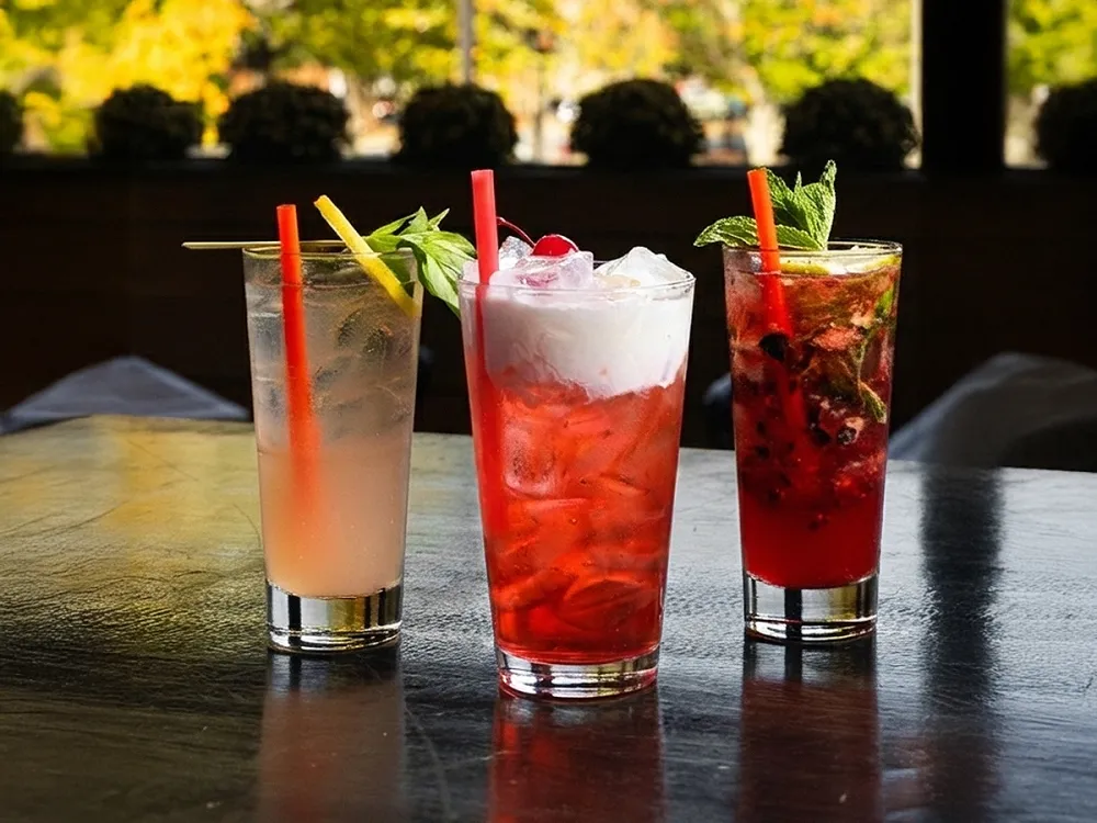 Three colorful iced cocktails with garnishes and straws on a dark reflective table with blurred greenery background