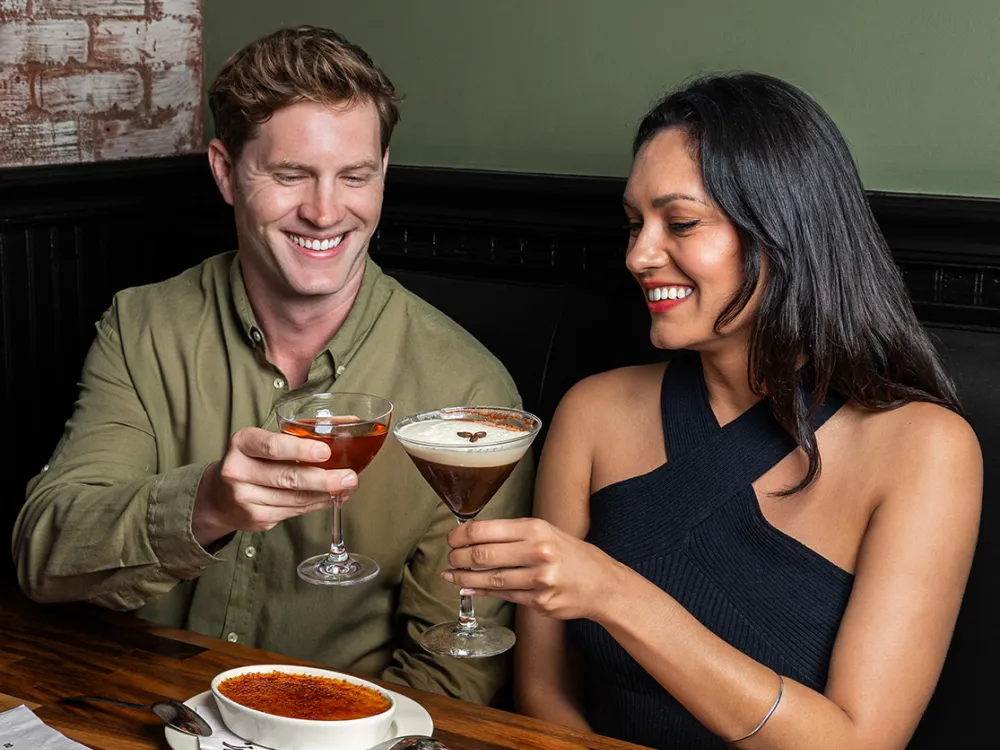 Smiling man and woman clinking cocktail glasses while seated at a wooden table with creme brulee dessert.