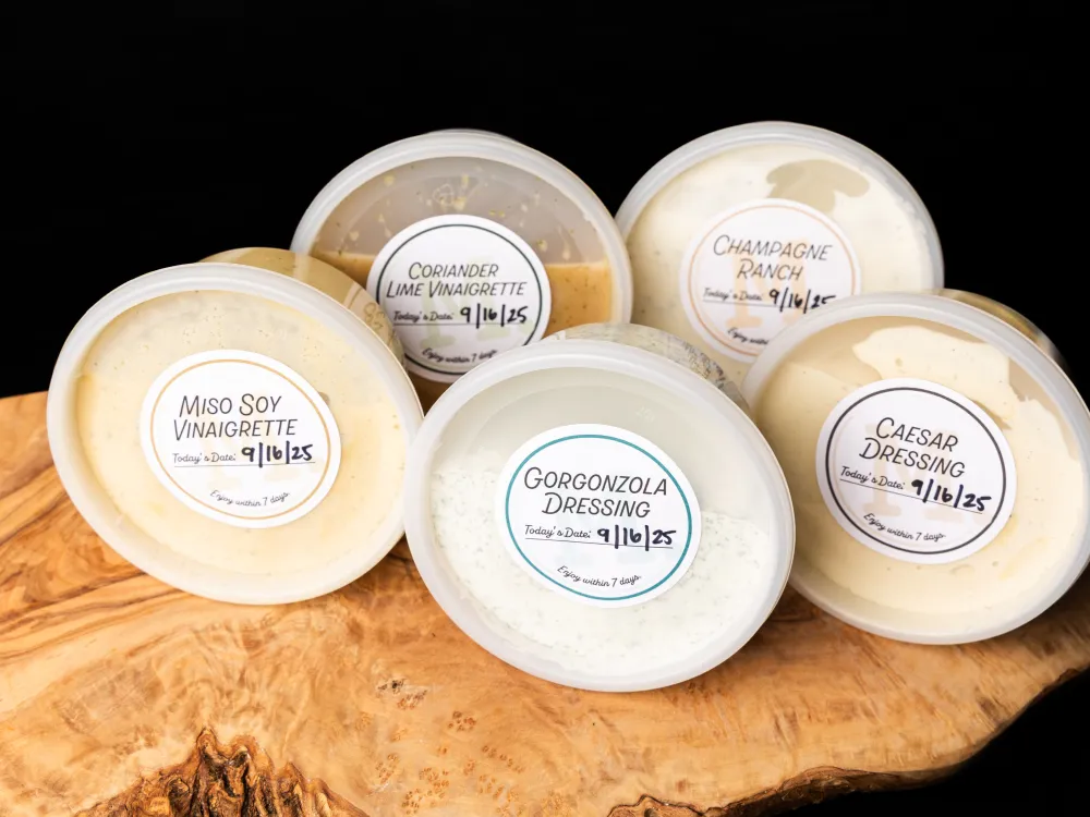 Five plastic containers of various salad dressings and vinaigrettes on a wooden surface against black background.