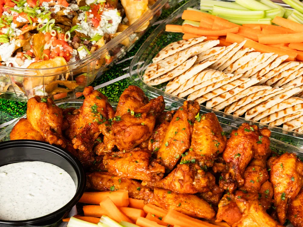 Spicy chicken wings with ranch dip, carrot sticks, celery, grilled flatbreads, and loaded nachos in trays.