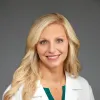 Smiling female doctor with long blonde hair wearing a white Pinnacle Orthopaedics coat and green scrub top.