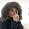 Woman in a winter coat with a fur hood warming her hands indoors near a thermostat.