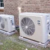 Two outdoor mini-split air conditioning units installed on concrete slabs beside a brick house wall with greenery around.
