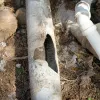 Broken underground concrete pipe with large hole next to intact white plastic pipes on soil