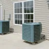 Two outdoor air conditioning units installed on a concrete patio beside a beige house wall with a window and door.