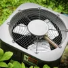 Outdoor air conditioning unit surrounded by green plants, showing the metal fan and protective grille.