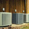 Three outdoor air conditioning units installed on concrete pad beside wooden wall in natural setting