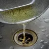 Used cooking oil being poured from a pan into a kitchen sink drain with water droplets around.
