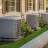 Three outdoor air conditioning units installed beside a beige house with green landscaping and stone bases.
