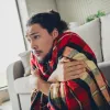 Man wrapped in a red plaid blanket sitting on the floor, looking cold and shivering indoors by the couch.