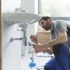 Plumber fixing a bathroom sink pipe using wrenches, with tools and plunger nearby in a modern bathroom.