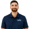 Man wearing navy blue polo with PV logo, beige pants, brown belt, and brown sneakers standing with hands in pockets.