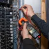 Electrician using clamp meter to test voltage in residential circuit breaker panel with multiple wires.