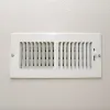 White rectangular air vent with adjustable louvers mounted on a beige wall with visible screws.