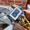 Hands using a digital manifold gauge to check pressure on an outdoor air conditioning unit with a fan.