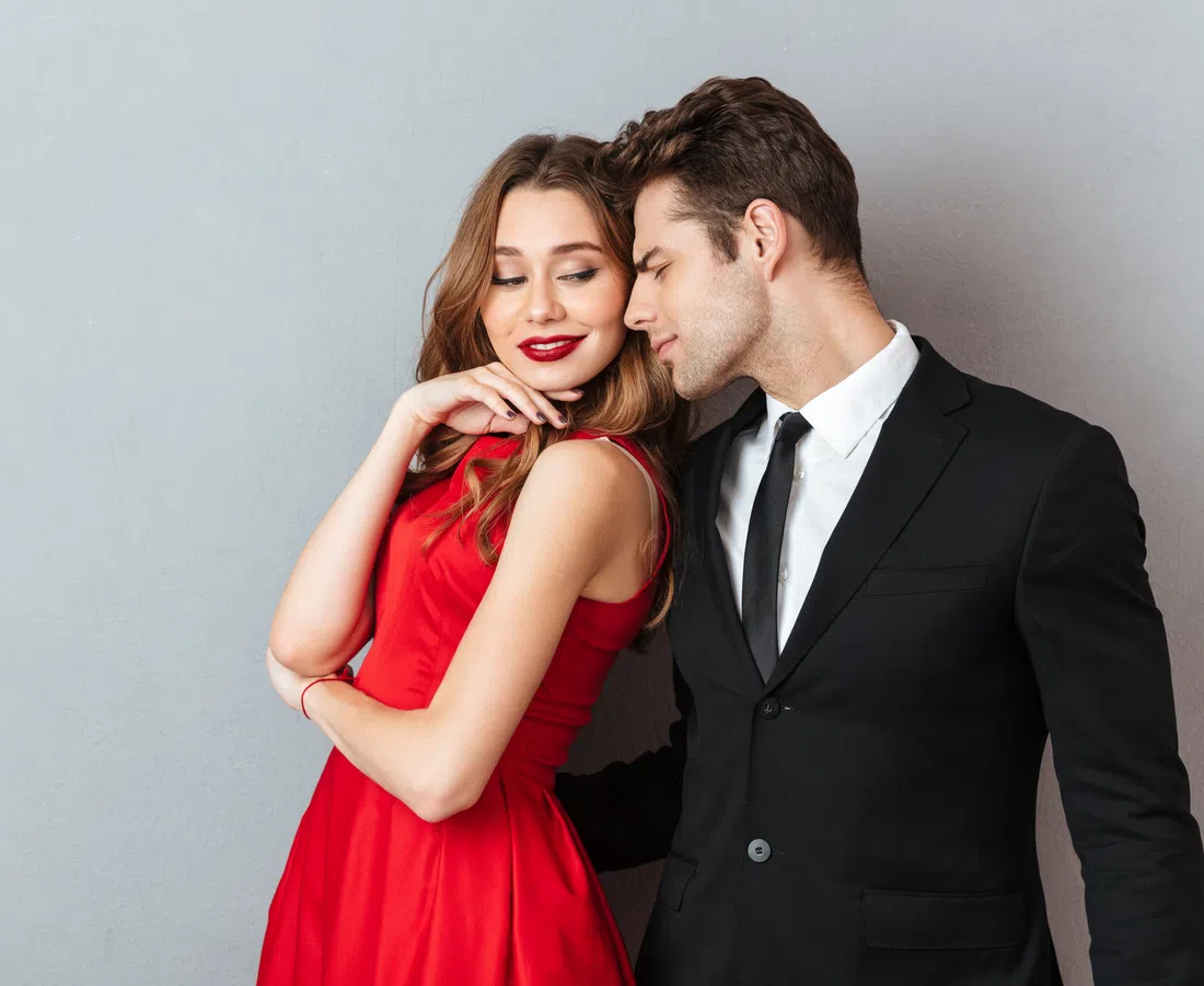 Stylish couple with woman in red dress and man in black suit posing intimately against gray background.