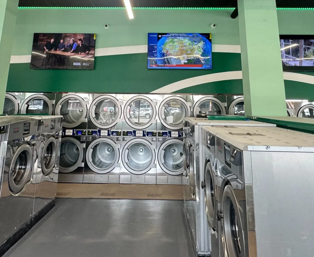 Interior of a modern laundromat with rows of stainless steel washing machines, green walls, and ceiling TVs.