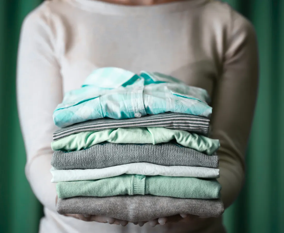 Person holding a neatly folded stack of pastel and neutral colored clothes with a green curtain background