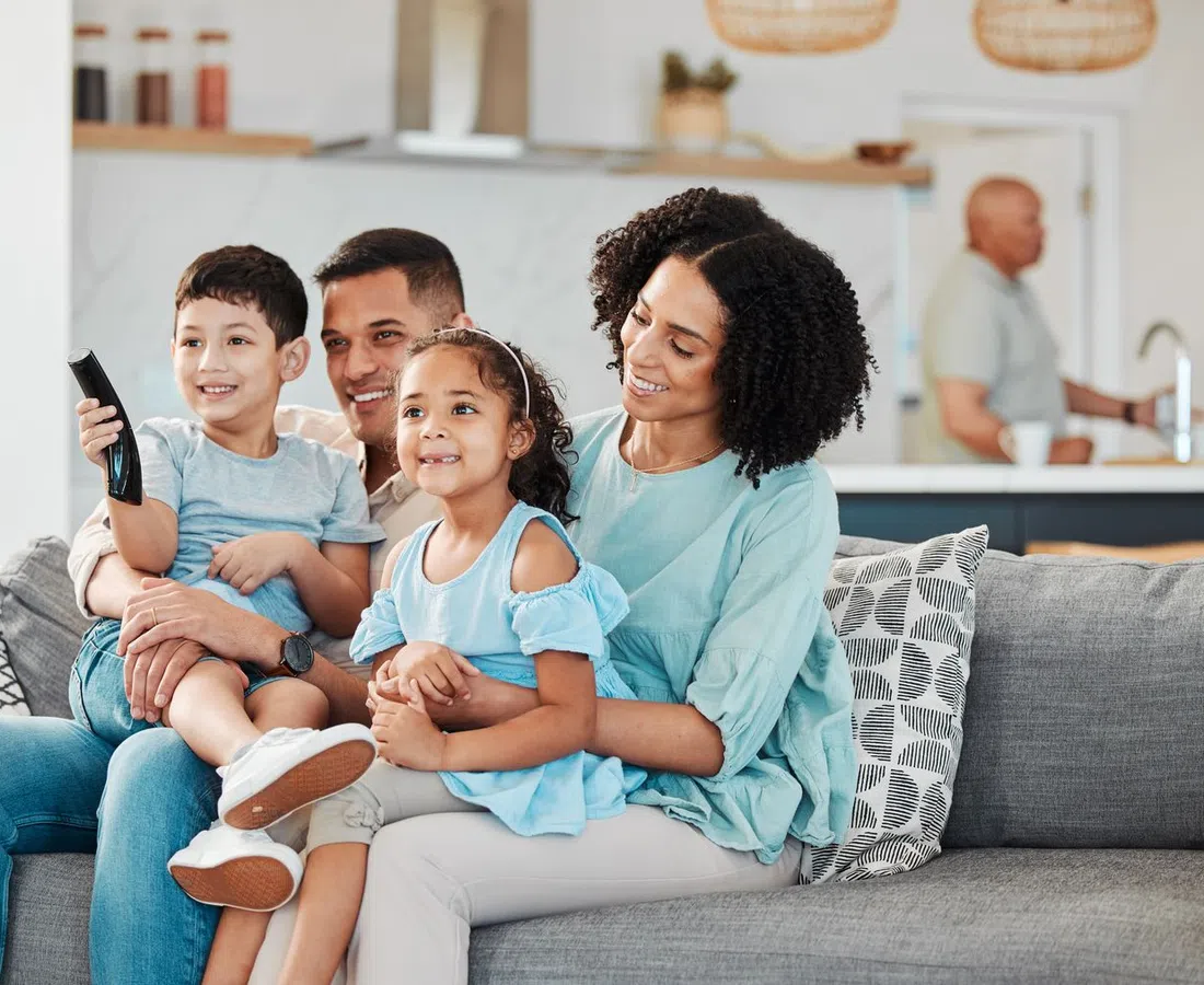 Happy family of four sitting on couch watching TV together in a cozy modern living room.