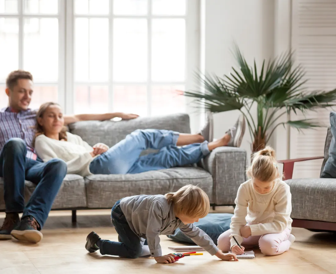 Parents relax on a couch while their children draw and play on the floor in a bright living room.