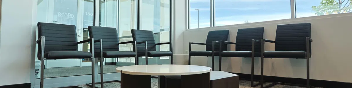 Modern waiting area with black chairs around a low round table on a gray carpet near large windows.