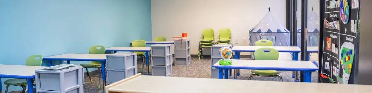 Bright and organized classroom with colorful chairs, desks, storage bins, and educational posters on the window.
