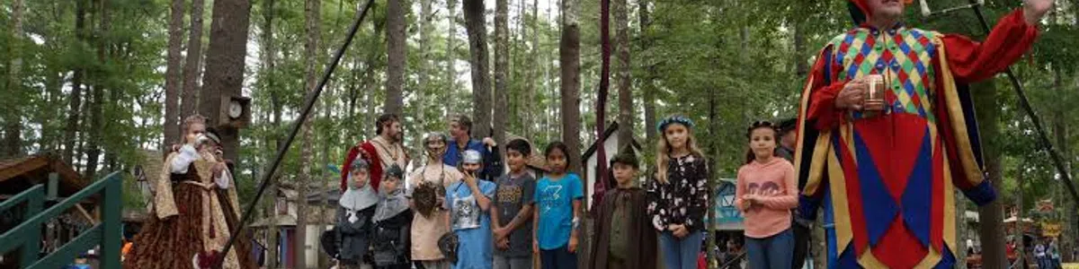 Children and adults in medieval costumes stand on an outdoor wooden stage surrounded by trees during a performance.