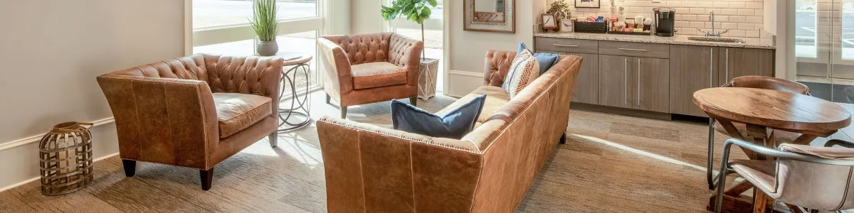 Bright modern lounge with leather sofas, circular LED ceiling light, kitchenette, and large windows providing natural light.