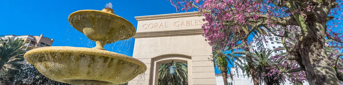 Coral Gables arch with water fountain and blooming pink tree under a clear blue sky in Florida cityscape.