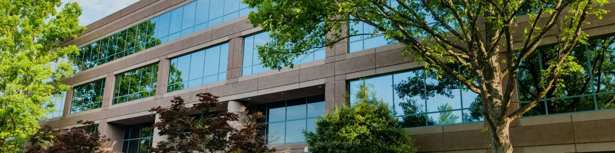 Modern office building with large glass windows surrounded by lush green trees and landscaped plants in daytime.