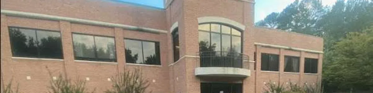 Two-story red brick office building with large windows and surrounding trees in a parking lot area.