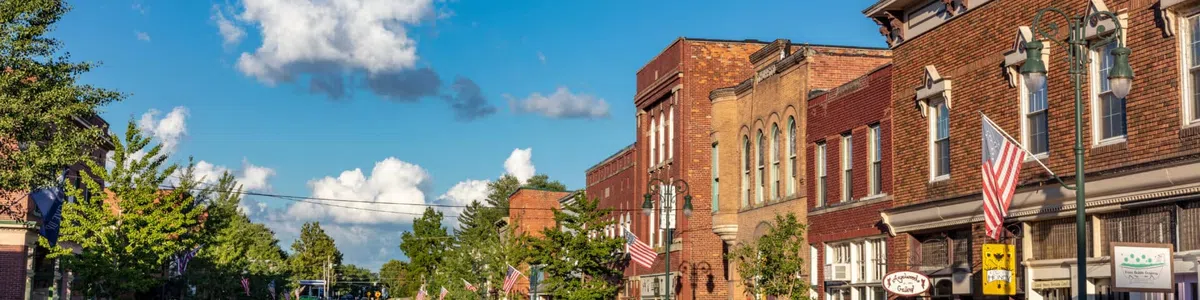 Sunny day view of a small town street lined with brick buildings, parked cars, and American flags.