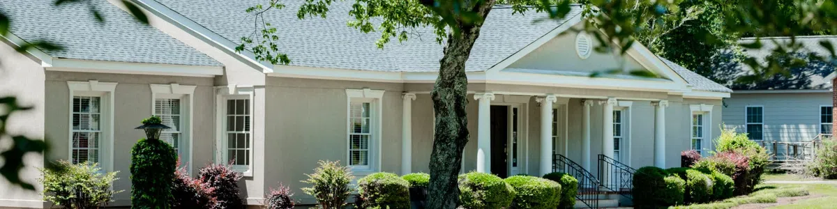 Single-story beige house with a blue-gray roof surrounded by greenery and shaded by trees