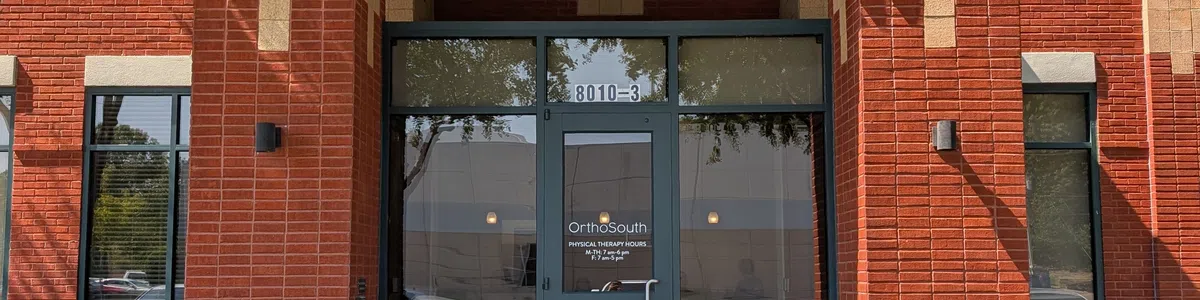 Entrance of OrthoSouth clinic with green signage, red brick walls, glass door, and surrounding greenery under blue sky.